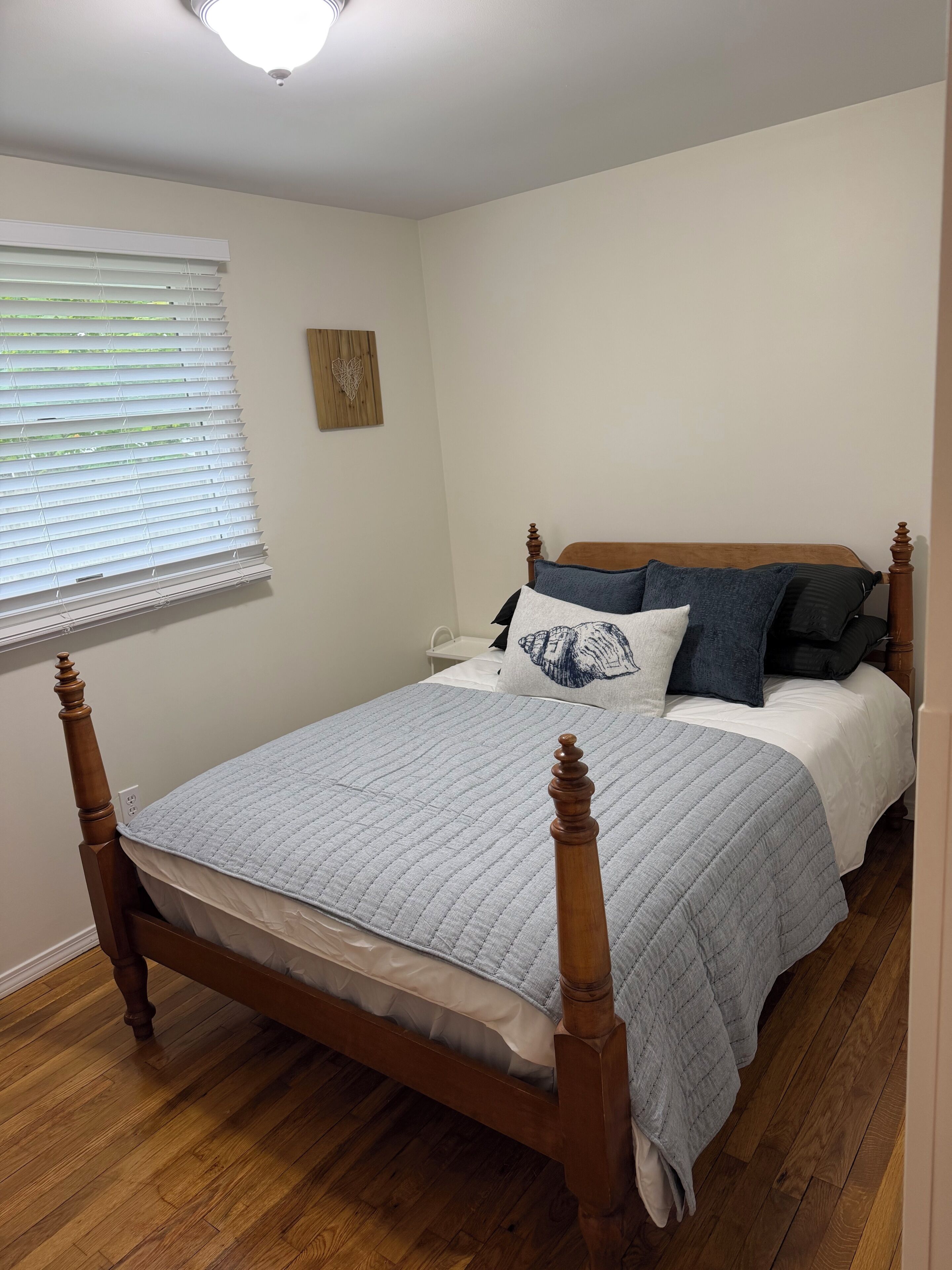 Une chambre avec un lit en bois, des murs blancs, une fenêtre avec des stores et une petite œuvre d’art en bois accrochée au mur.