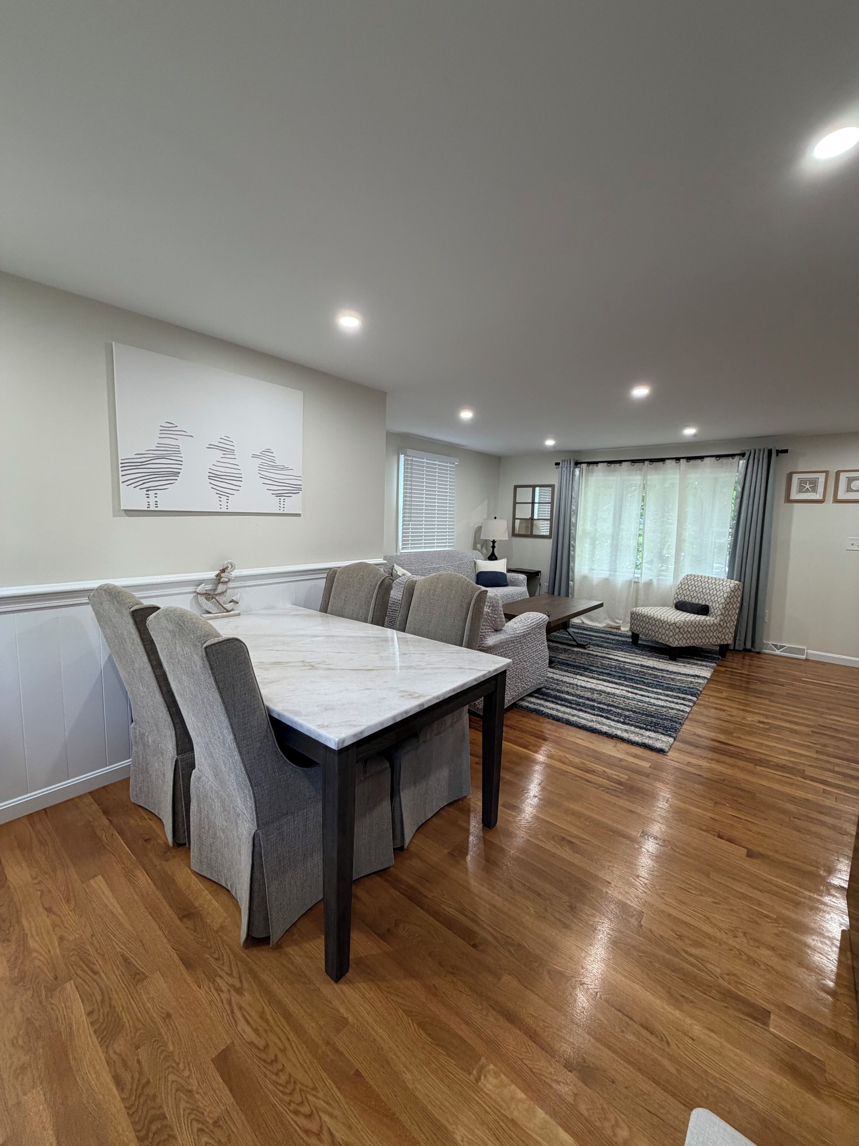 A modern living room with a dining area, wooden flooring, and a large window with curtains.