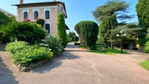 Una strada privata lastricata che conduce a un edificio con facciata rosa, immerso nel verde e alberato.