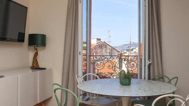 A room with a round table, chairs, a TV, and a view of buildings through a window. - Agréable et Cosy - Suquet - proche des plages (Cannes)