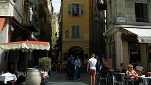 Une rue étroite bordée d’immeubles, avec des terrasses et des passants.