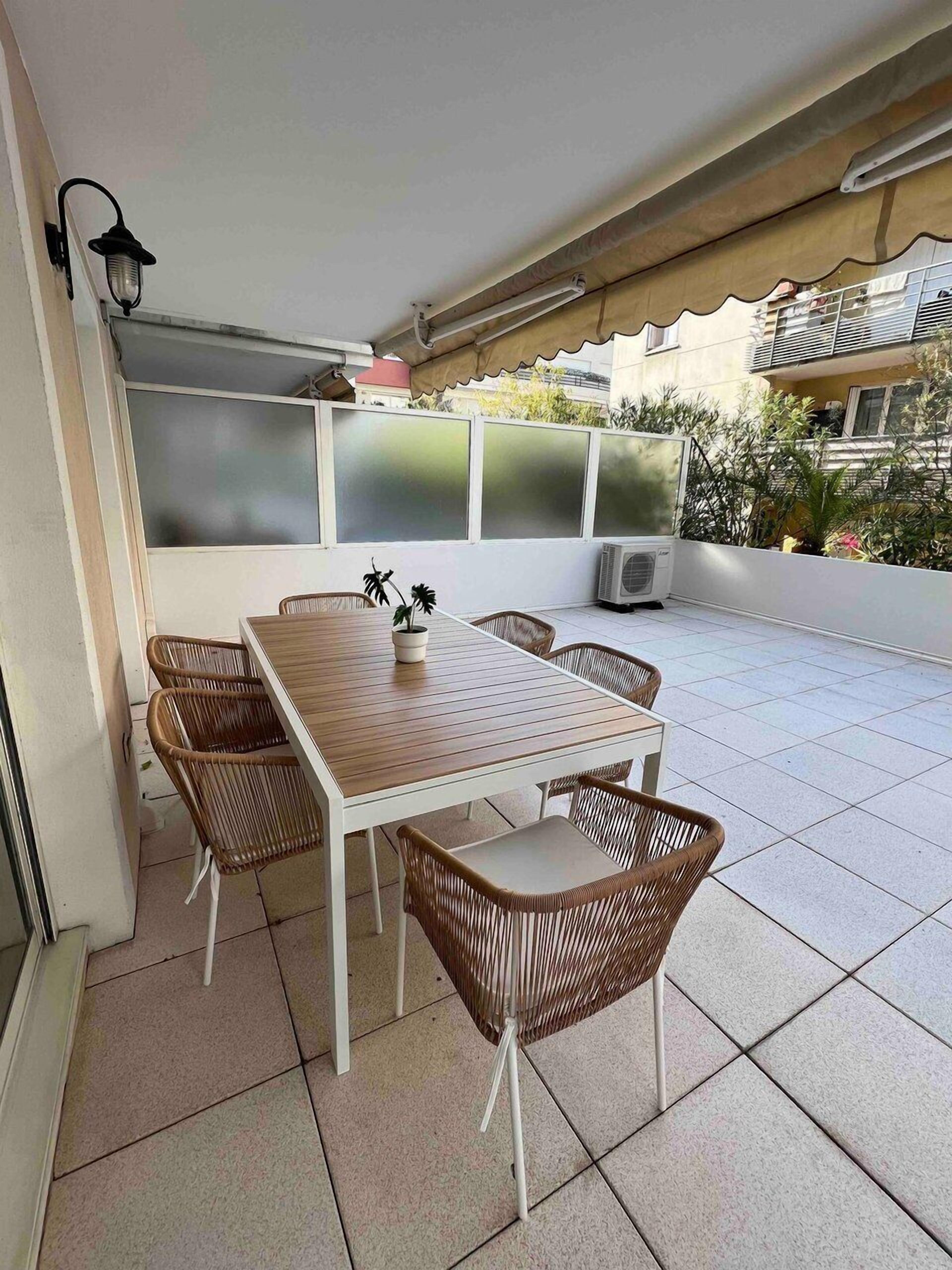 Eine Terrasse mit einem weißen Tisch, Korbstühlen, einer Klimaanlage und Blick auf ein Wohnhaus.