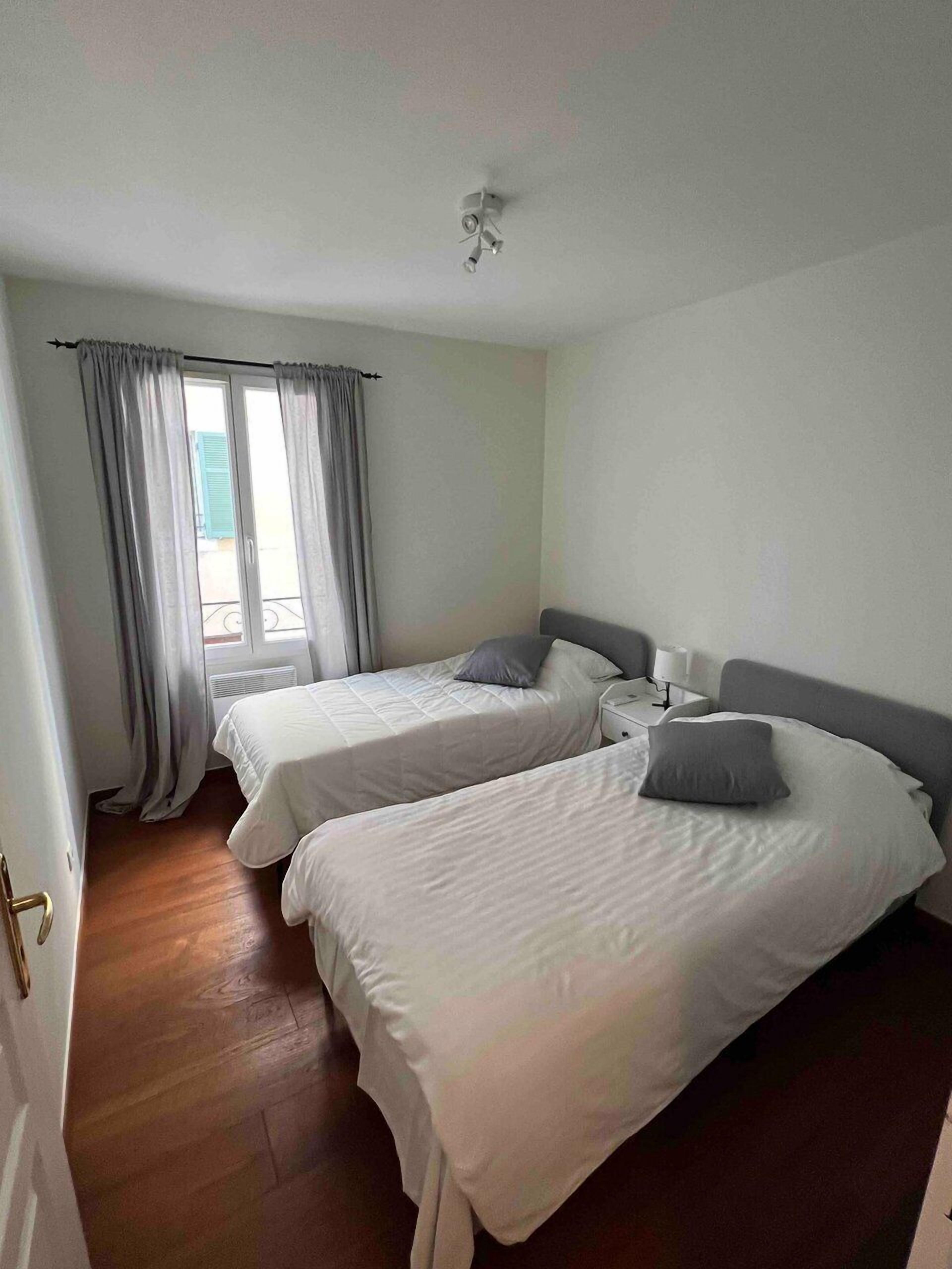 A hotel room with two beds, a window with curtains, a bedside table with a lamp, and a wooden floor.