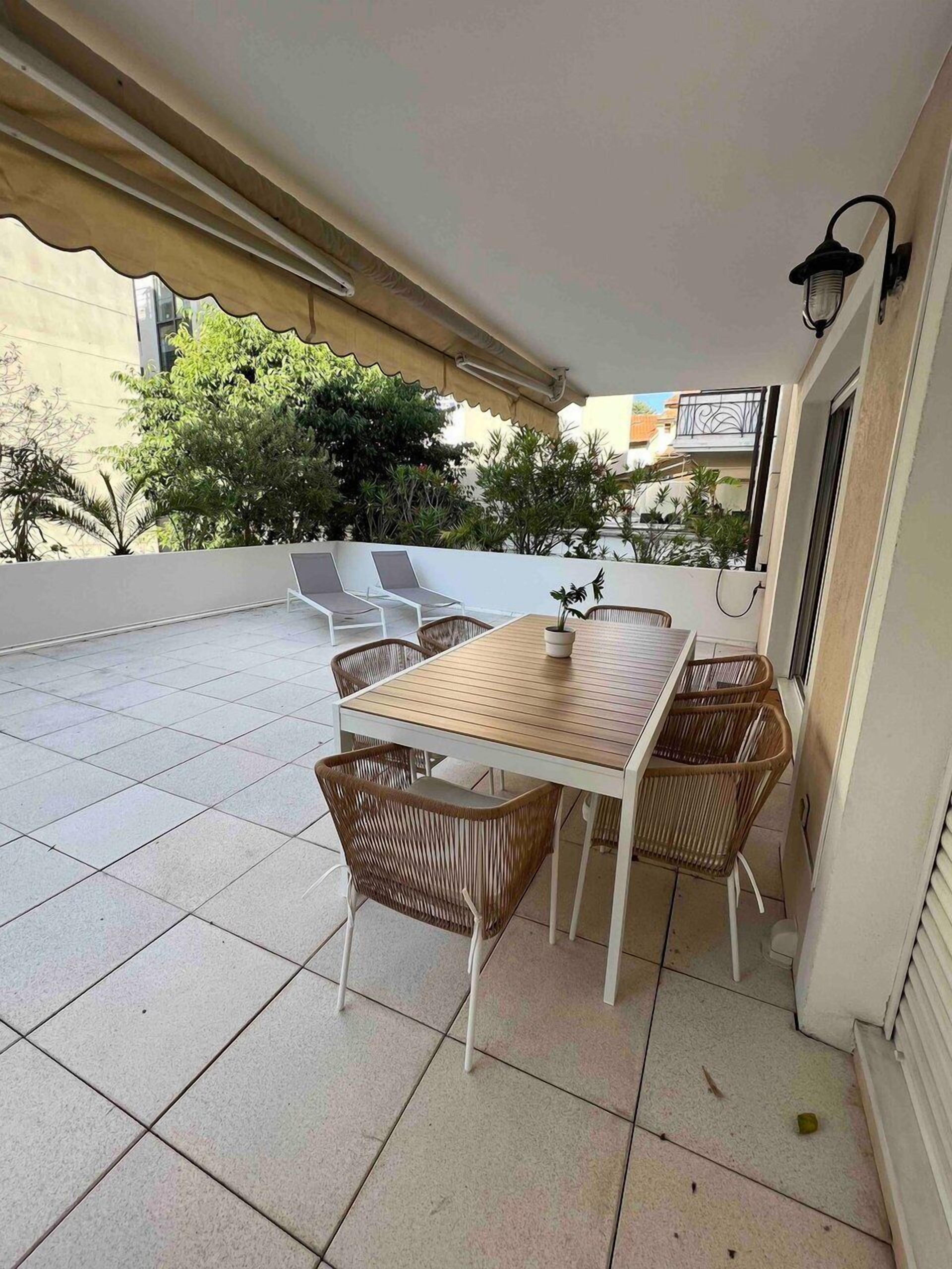 Une terrasse couverte avec une table et des chaises, un sol carrelé et une vue sur les bâtiments et la végétation environnants.