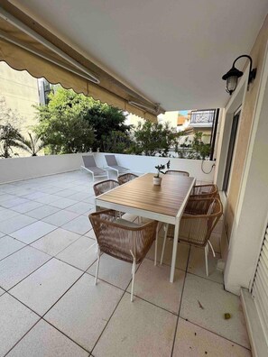 Eine überdachte Terrasse mit Tisch und Stühlen, einem gefliesten Boden und Blick auf umliegende Gebäude und Grünflächen.