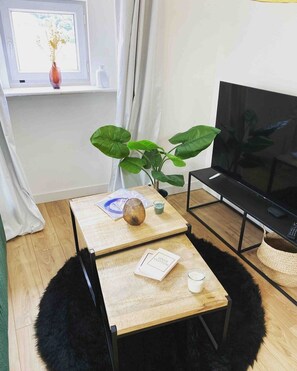 Ein Zimmer mit einem Holztisch, einem Fernseher, einer Pflanze und einem Fenster mit Vorhängen.