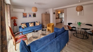 Uma sala de estar com um sofá azul, uma poltrona amarela e um espaço de jantar com uma mesa e cadeiras brancas.