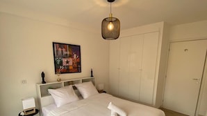 Uma cama bem arrumada com uma toalha enrolada, uma mesinha de cabeceira com uma caixa, um quadro na parede e um lustre no teto.