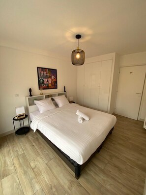 Ein ordentlich bezogenes Bett mit einem zusammengerollten Handtuch, ein Nachttisch mit einer Schachtel, ein an der Wand hängendes Bild und eine Deckenleuchte.
