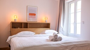 A neatly made bed with white linens, two bedside lamps, a framed abstract artwork, and a window with curtains. - Agréable et Cosy - Suquet - proche des plages (Cannes)