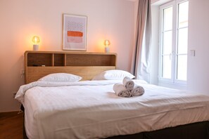 A neatly made bed with white linens, two bedside lamps, a framed abstract artwork, and a window with curtains. - Agréable et Cosy - Suquet - proche des plages (Cannes)