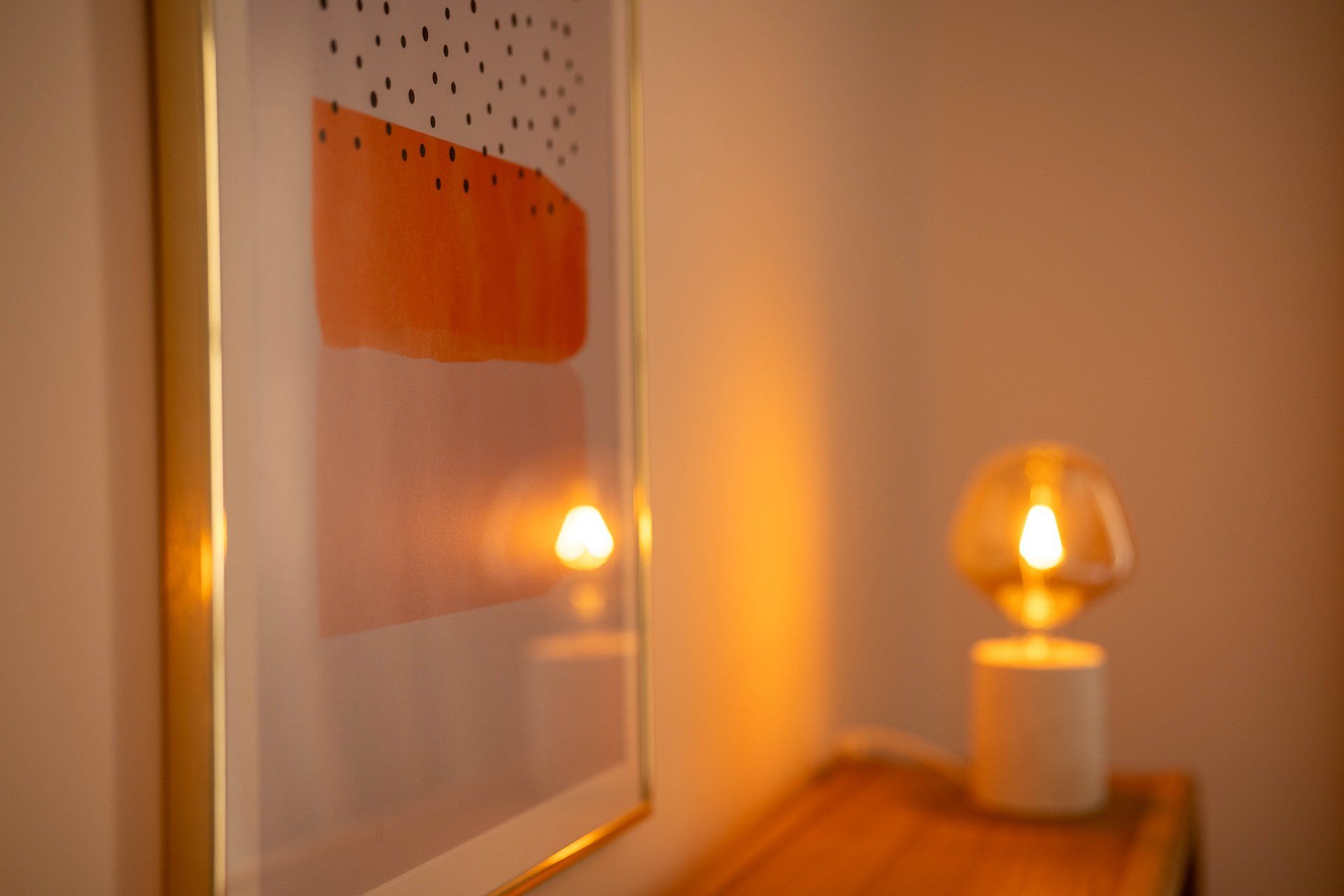Ett inramat konstverk med ett prickmönster och en abstrakt del i varma toner, en bordslampa och en träyta.