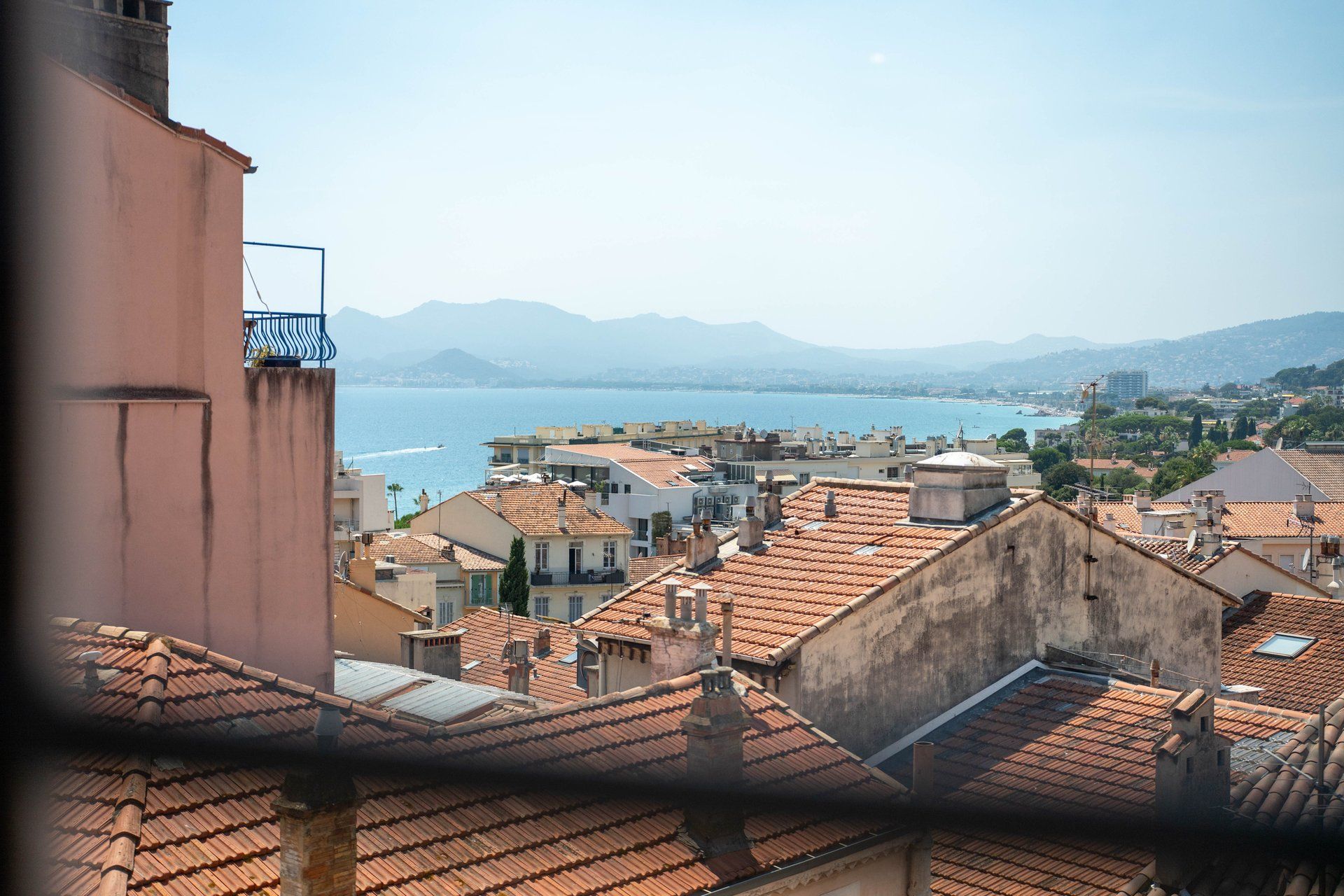 Un paisaje urbano costero con edificios de tejados de terracota, vistas claras al mar y montañas en la distancia.