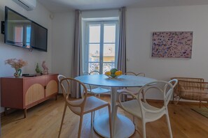 A dining area with a round table, white chairs, a fruit bowl, a pink cabinet, and a television on the wall. - Agréable et Cosy - Suquet - proche des plages (Cannes)