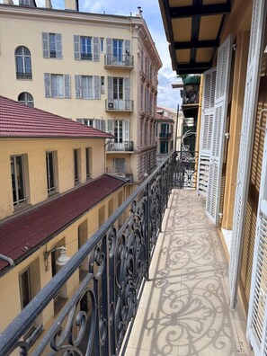 Ein Balkon mit aufwendiger Metallgitterarbeiten, angrenzende Gebäude mit zahlreichen Fenstern und Balkonen sowie ein rotes Ziegeldach.