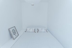 Ein schlicht eingerichtetes Zimmer mit einem Einzelbett, einem gerahmten Bild an der Wand und einer Deckenleuchte.