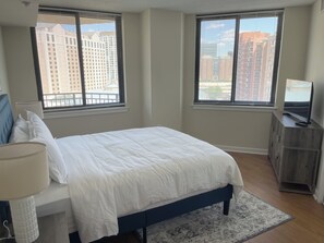Ein Schlafzimmer mit einem Bett, einem Fernseher auf einer Holzkommode und zwei Fenstern mit Blick auf die Stadt.