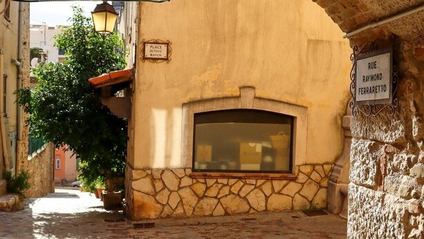 An arched passageway with a stone wall, a window, and a sign indicating 'RUE RAYMOND FERRARETTO.'