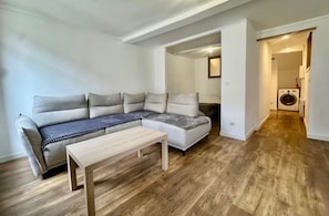 Ein modernes Wohnzimmer mit einem Ecksofa, einem Holztisch und einem durch eine offene Tür einsehbarer Waschbereich.