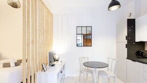 Een moderne, minimalistische kamer met een bed, een bureau met een computer, een ronde tafel met stoelen en een keukenhoek.