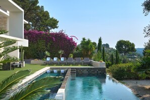 Una piscina amb gandules, un edifici modern i vegetació abundant.