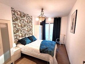 Ein Schlafzimmer mit Bett, Nachttisch, Schminktisch, einem Fenster mit Vorhängen, einer Heizung und einer Kandelaberleuchte.