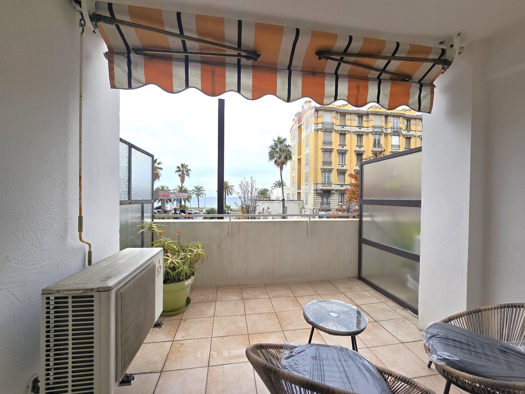 A balcony with a striped awning, a potted plant, a table, and chairs.