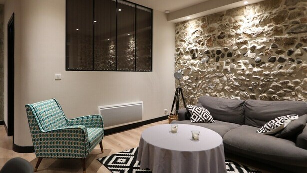 A modern living room with a stone wall, a large window, a patterned armchair, a grey sofa, and a round coffee table. - Superbe Appartement moderne au coeur du Vieux Nice (Nice)