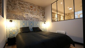 A bedroom with a stone wall, a bed with black bedding, a small bedside table, and a mirror on the wall. - Superbe Appartement moderne au coeur du Vieux Nice (Nice)