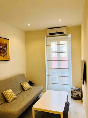 Una sala de estar con sofá, una mesa de centro, televisor, equipo de aire acondicionado y ventana con persianas.