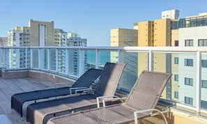 A rooftop terrace with two wicker lounge chairs, a glass balustrade, and a cityscape view. - Apartamento verão na praia (Vila Velha)