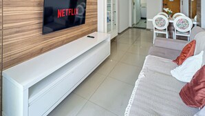 Una sala de estar moderna con televisor de pantalla plana, pared de paneles de madera, mueble de entretenimiento blanco y un área de comedor con sillas y una mesa.