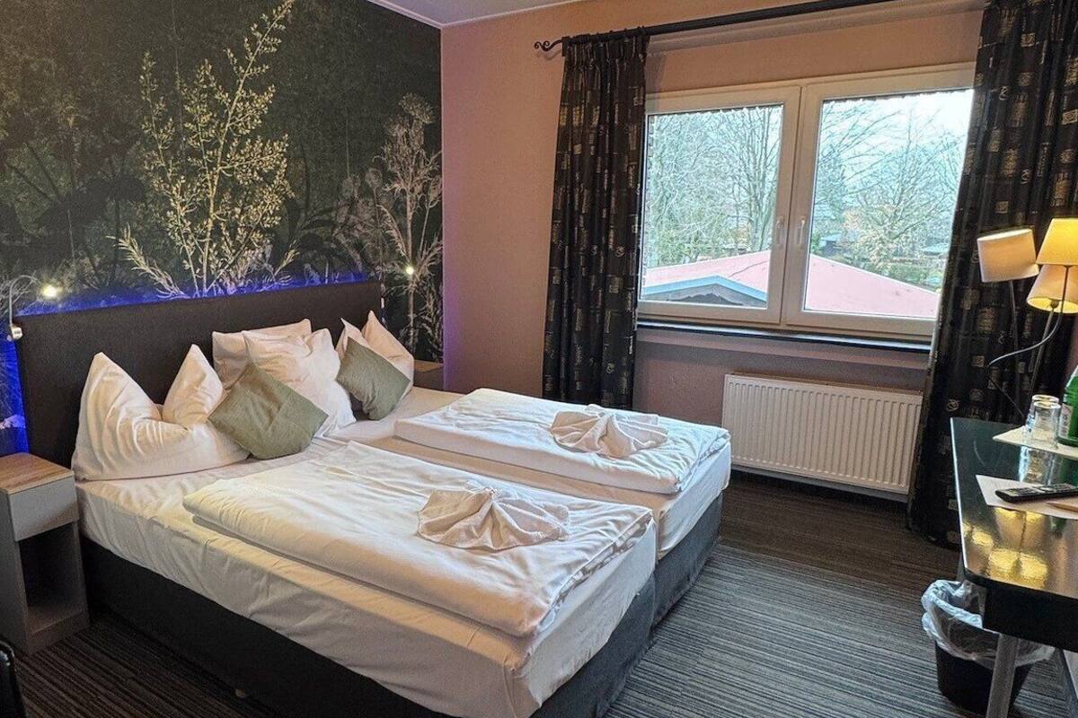 A hotel room with a bed, a nightstand, a table with a lamp, a view of trees through the window, and a radiator.