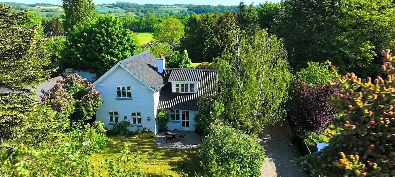 Une maison blanche, coiffée d’un toit gris, est entourée d’une végétation luxuriante et d’arbres.