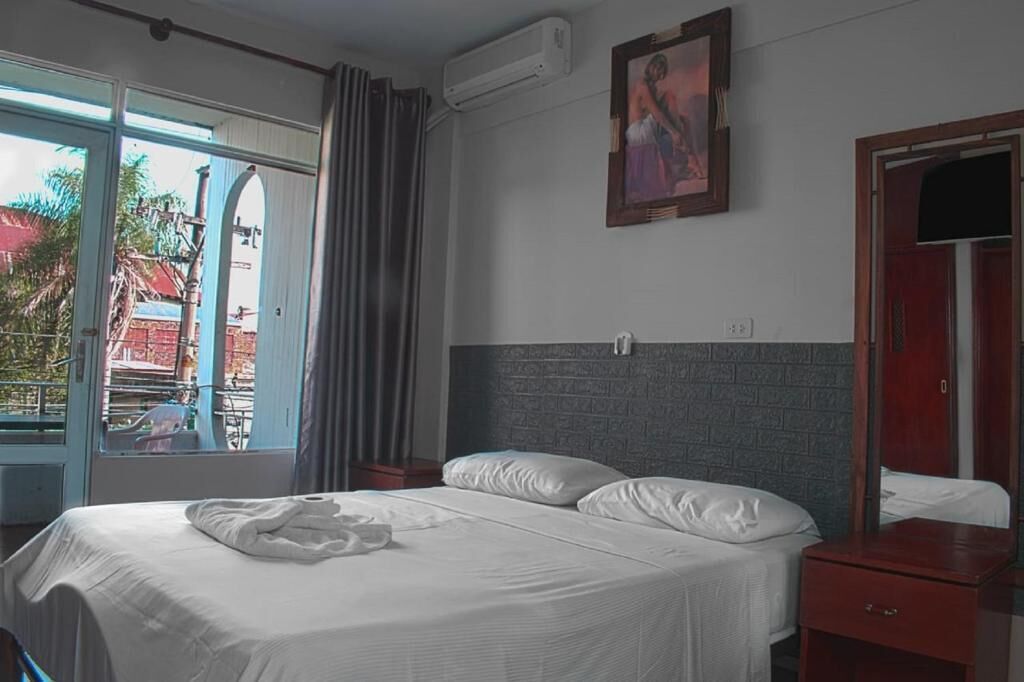 A hotel room with a bed, a nightstand, a wooden wardrobe, a window with curtains, and an air conditioning unit.