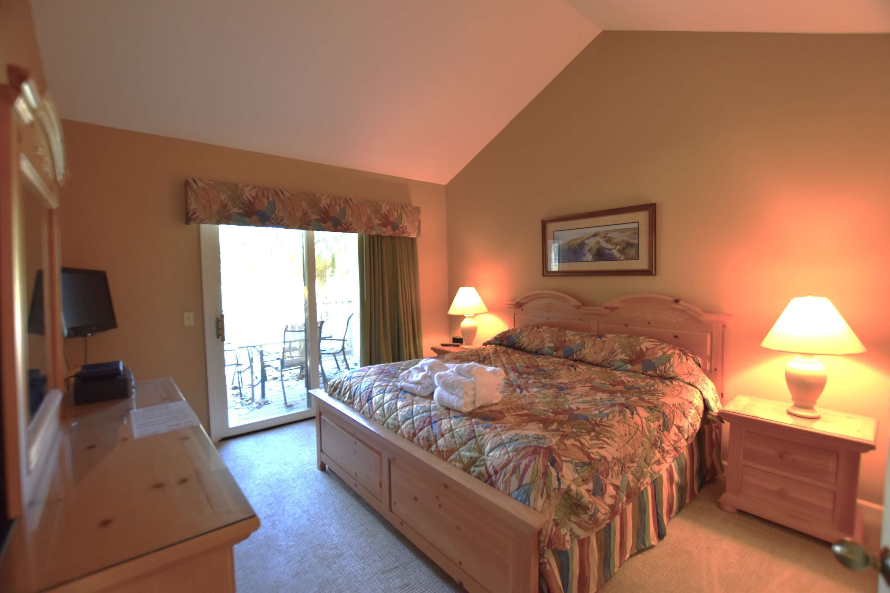 A bedroom with a bed, two bedside lamps, a desk with a lamp, a chair, and a sliding glass door leading to an outdoor area.