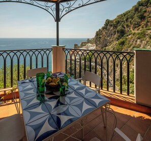 A table with a geometric pattern, a basket of lemons, and green glasses on a balcony overlooking a coastal landscape. - Seaside house with terrace in Atrani (Ravello)