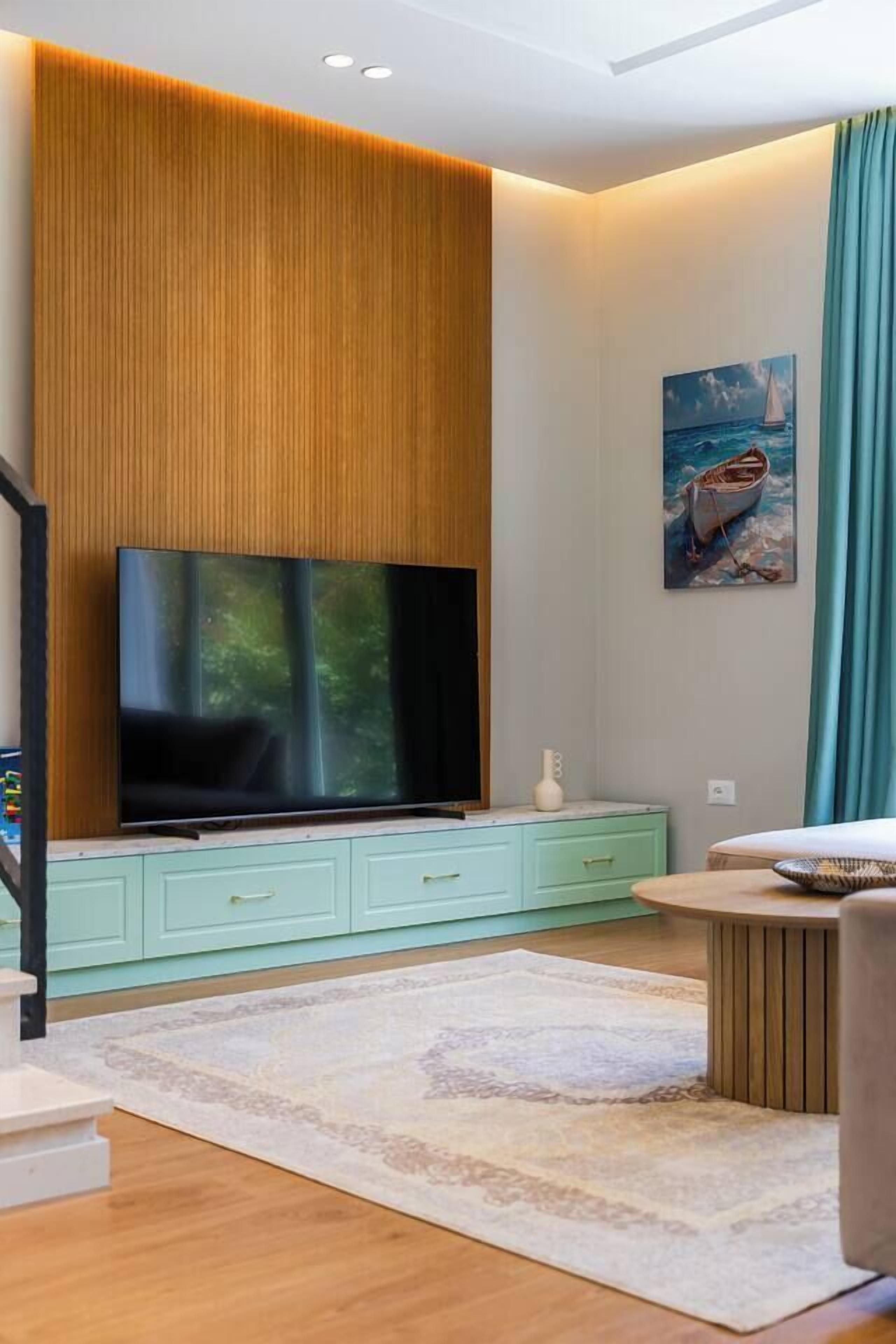 A modern living room with a flat-screen TV, light green cabinetry, a patterned rug, and a painting of a boat on the wall.