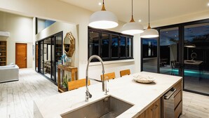 Une cuisine moderne dotée d’un grand îlot central, de luminaires suspendus et offrant une vue sur la piscine.