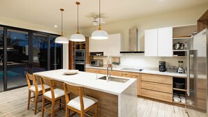 Une cuisine moderne avec un îlot central, des chaises en bois, des luminaires suspendus et une vue sur une piscine.