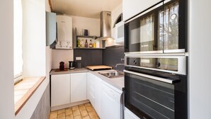 A compact kitchen with a dishwasher, oven, and a window with curtains. - Les Escapades - à 10min en voiture de la plage (Saint-Quay-Perros)
