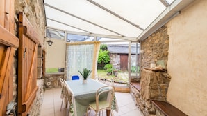 A covered patio with a table and chairs, stone walls, and a view of the garden. - Les Escapades - à 10min en voiture de la plage (Saint-Quay-Perros)
