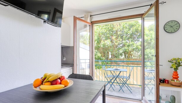 Une pièce comprenant une table à manger, un panier de fruits, une porte-fenêtre coulissante donnant sur un espace extérieur avec une table et des chaises, et une horloge au mur.