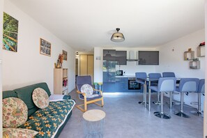 A modern living room with a sofa, a round coffee table, a dining area with bar stools, a kitchen with a microwave, and a ceiling light fixture. - L’Escapade du Ponant - À 3 km de la plage (Saint Raphael)