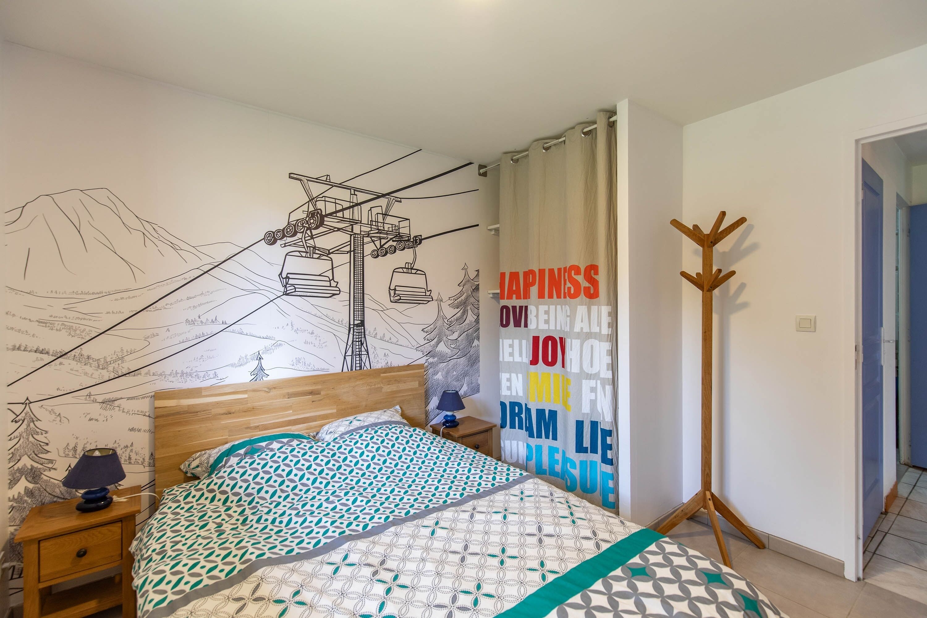 Une chambre à coucher comprenant un lit, une table de chevet, une lampe, un porte-manteau et un mur orné d’une illustration représentant un télésiège.