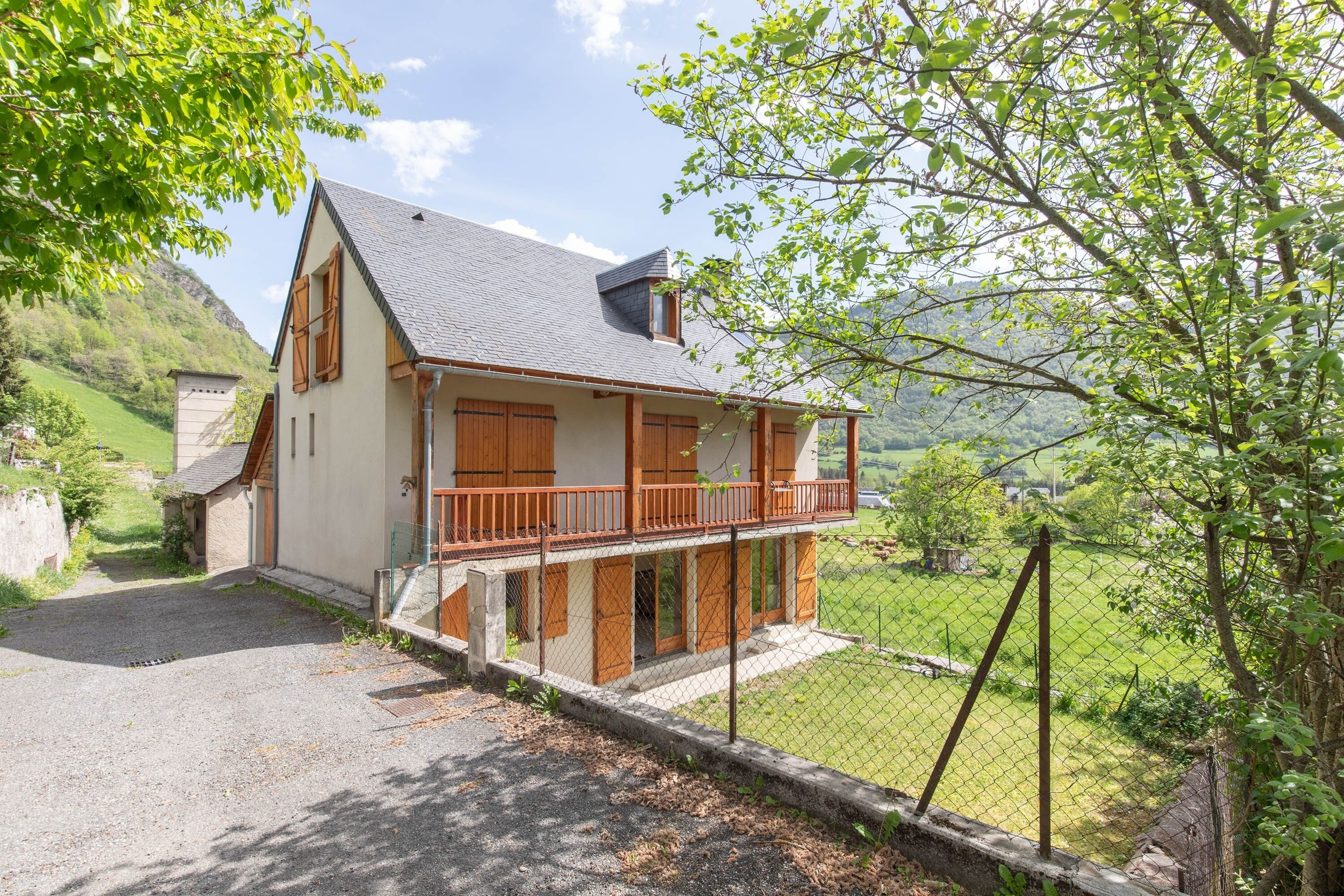 A two-story house with a balcony, a fenced yard, and a mountainous backdrop.