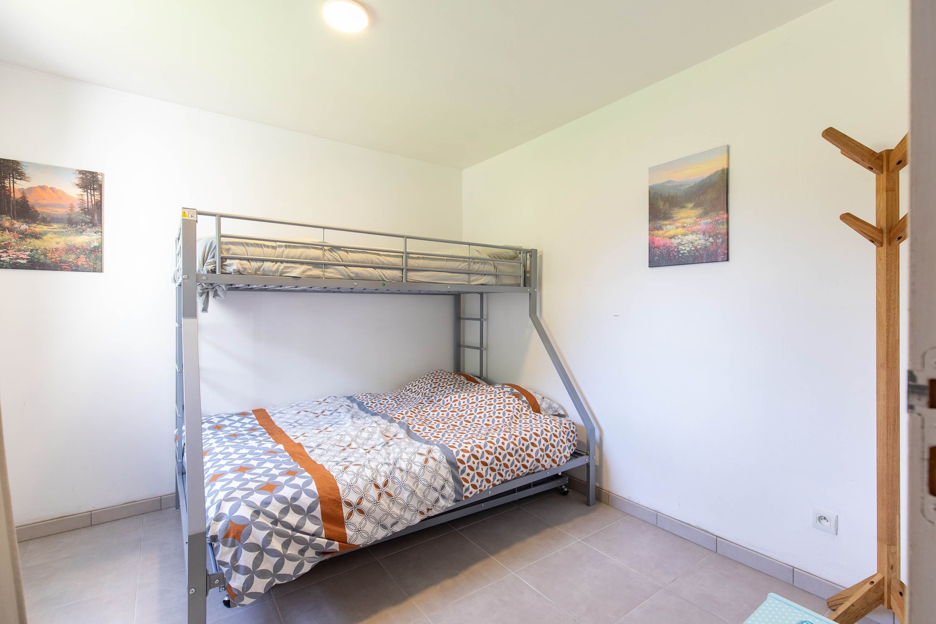 Una cama individual superpuesta con una colcha estampada, un perchero de madera y dos cuadros de paisajes en las paredes.