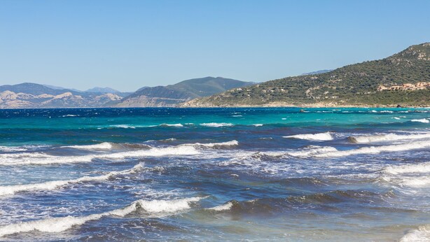 Un paysage côtier avec des vagues, un ciel dégagé et des montagnes à l’horizon.
