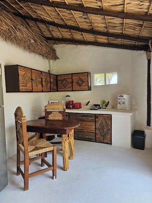 A rustic kitchen with a thatched roof, wooden cabinets, a dining table with chairs, and a window.