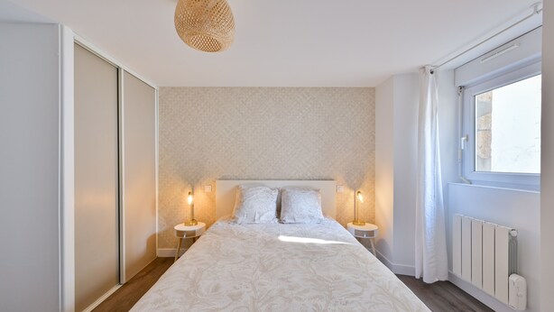 A bedroom with a large bed, two bedside tables with lamps, a ceiling light, a window with curtains, and a radiator. - L’Escale morbihannaise – Appt à 15 min de la plage (Muzillac)